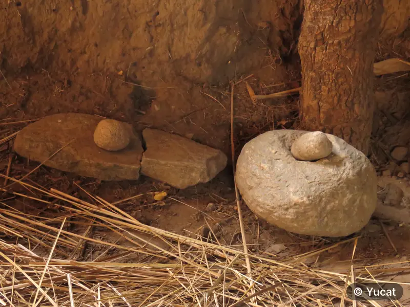 Dos morteros de piedra apoyados junto al muro de tierra de una casa-pozo, con paja en el piso.