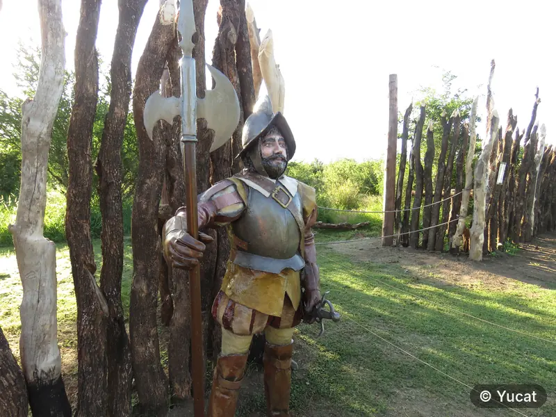 Escultura de un soldado español con armadura y alabarda, junto a la empalizada de troncos del fuerte.