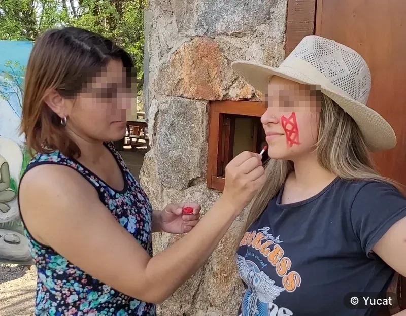 Una mujer pinta con un pincel un diseño rojo en la mejilla de una visitante, junto a un muro de piedra.